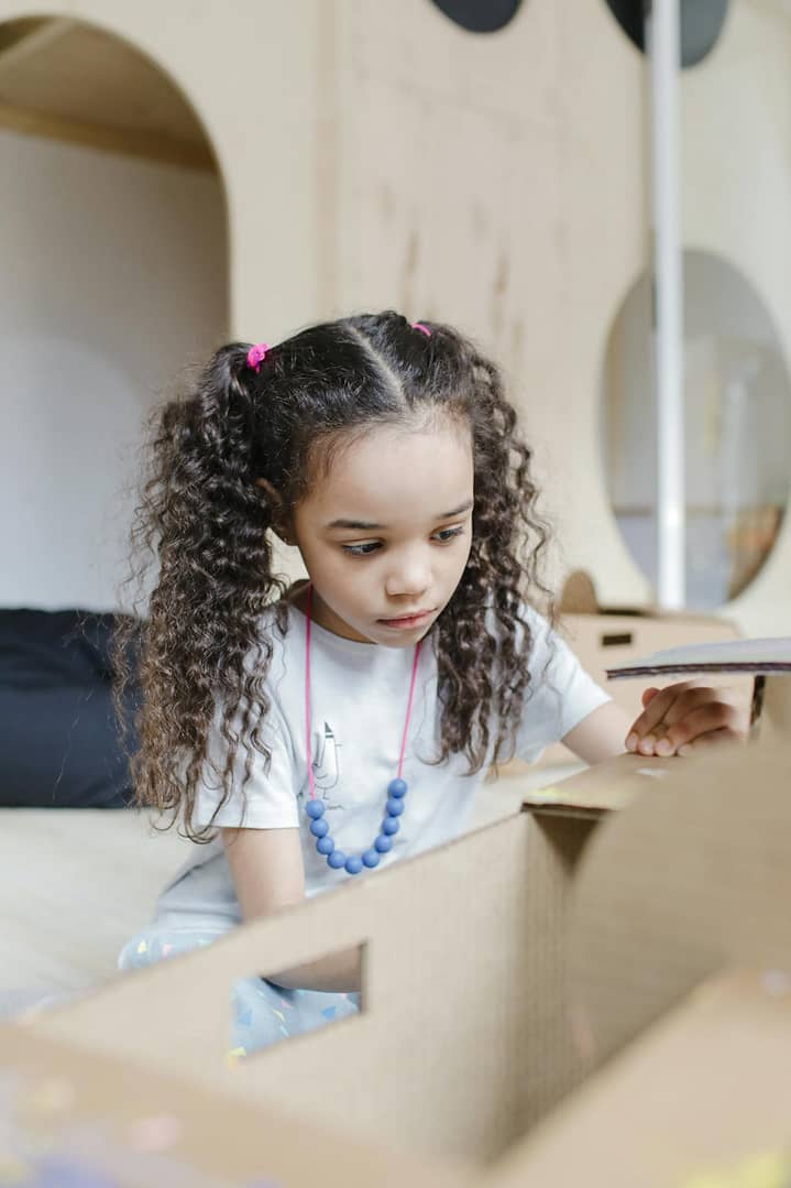 A young girl with curly hair playing creatively with a cardboard box indoors.